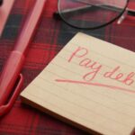 A handwritten note reading pay debt next to a pen and glasses, symbolizing a debt payoff plan