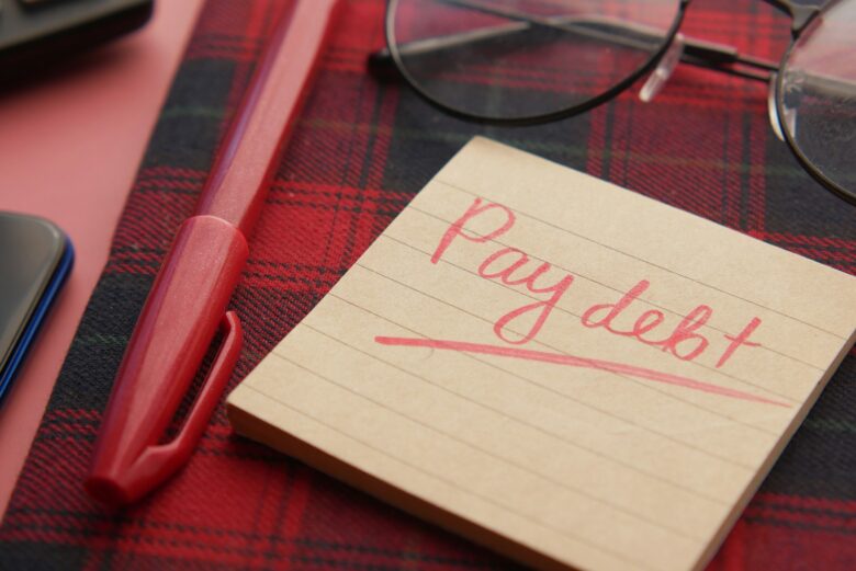 A handwritten note reading pay debt next to a pen and glasses, symbolizing a debt payoff plan